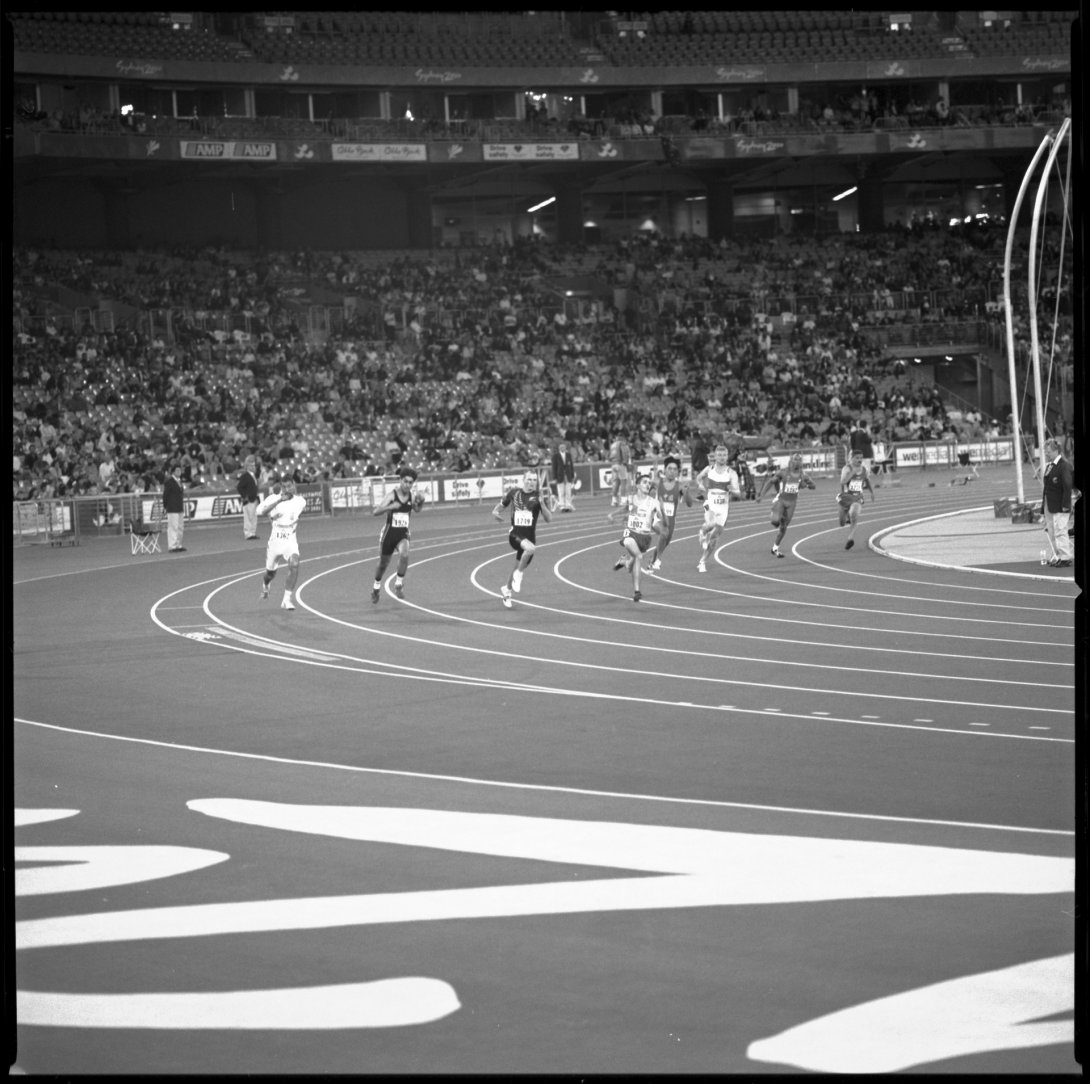 Eight men round the bend in a track running event. The stadium seating behind them is half full of spectators.