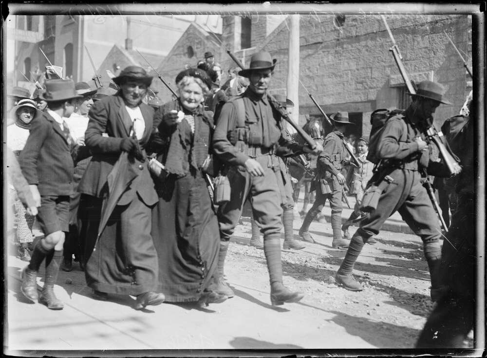 Men in military uniforms, with rifles over their shoulders, walk down an outdoor street surrounded by women and children. Two soldiers in the foreground are arm in arm with a woman