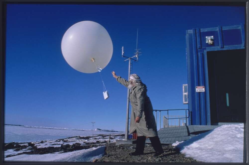 Meteorologist Neville Martin releases a large white weather balloon into a blue sky in Antarctica. the ground is snowy.