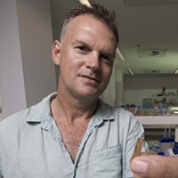 A portrait image of Professor Geoffrey Clark. He is wearing a light green/grey collared shirt. He is standing in an office. He holds a fragment of something in his fingers close to the camera