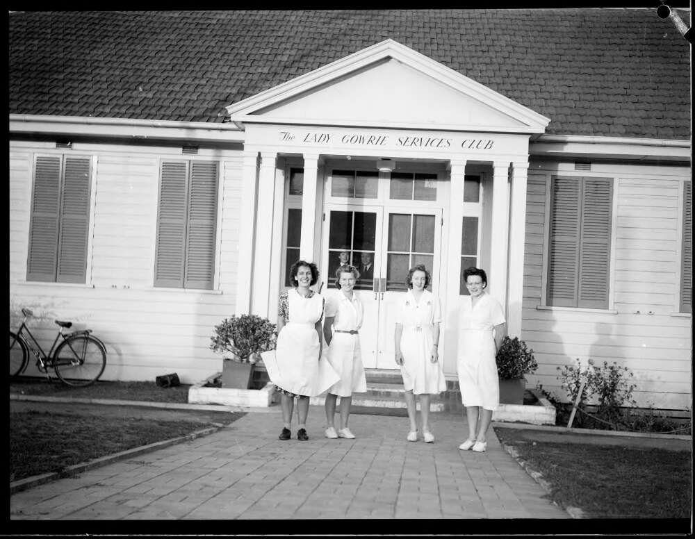 Four women in white dresses stand outside a large house
