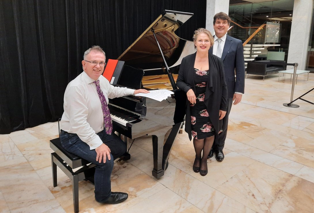 Three people stand around a baby grand piano. They are smiling at the camera.