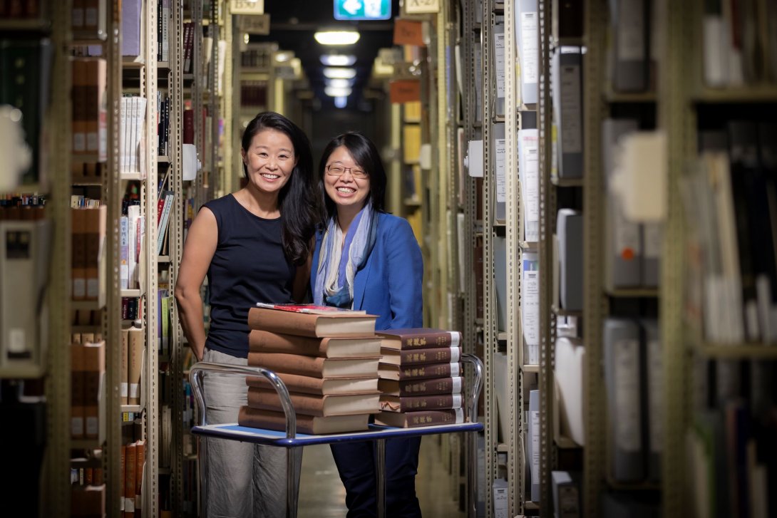 Two people are pictured amongst shelves of books. They have a trolley in front of them piled with stacks of leather-bound volumes.