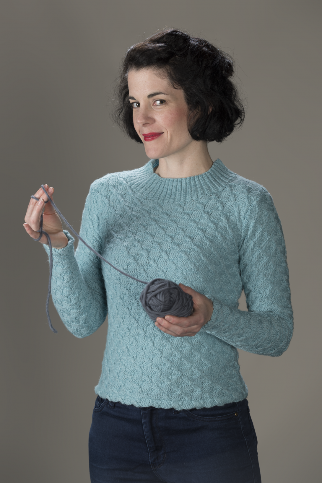 A woman in a light blue knitted jumper holds a ball of yarn in her left hand, with a loose thread held in the right.