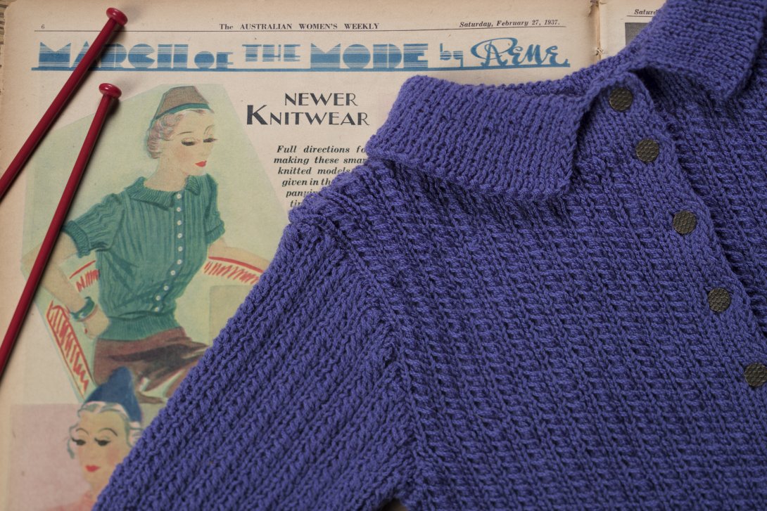 A knitted purple cardigan is laid over a vintage copy of the Australian Women's Weekly, containing an editorial about knitting