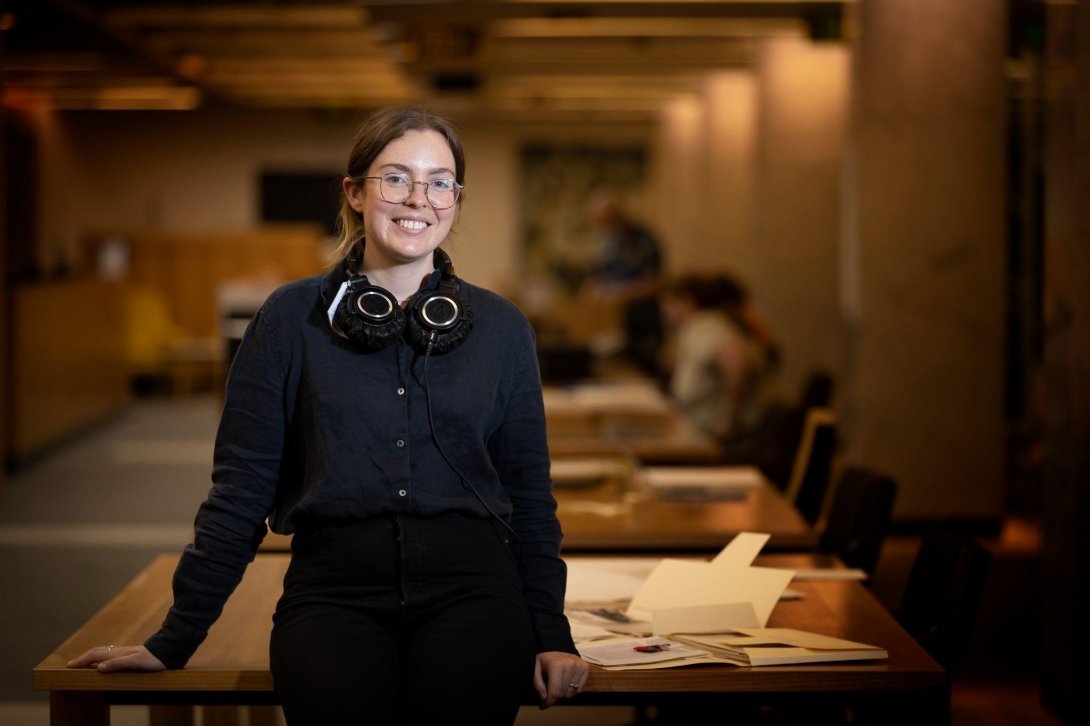 A scholar smiles at the camera. She is standing in the Library's special collections reading rooms - other people, desks and collection material are visible behind her.