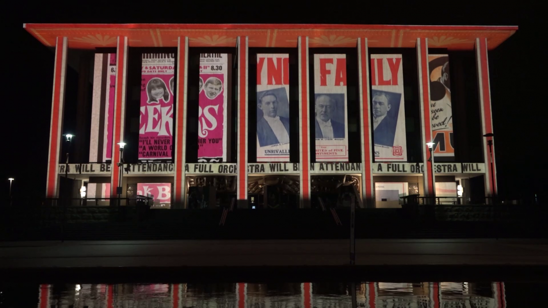 Exterior of the National Library building at light, illuminated with projections featuring playbills, theatre posters and printed programmes