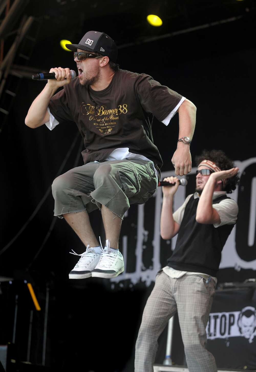 A man in a cap and sunglasses jumps in the air while singing into a microphone