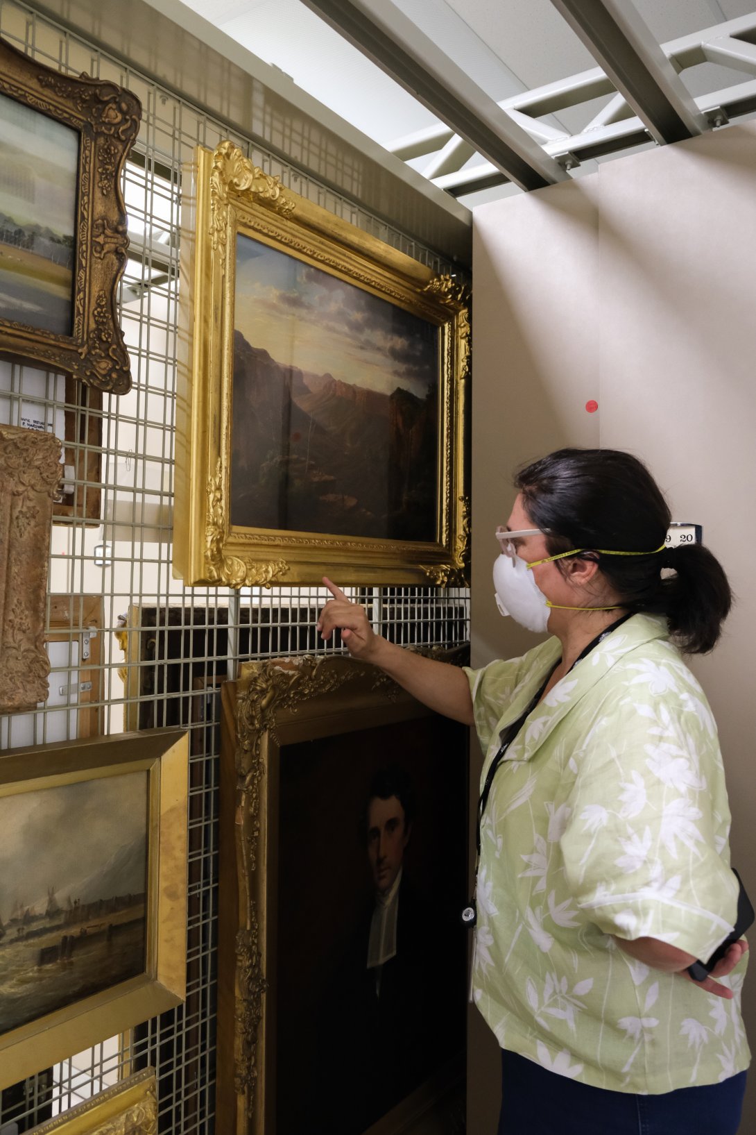 A woman in a white shirt and a face mask observes a painting in a gilded gold frame