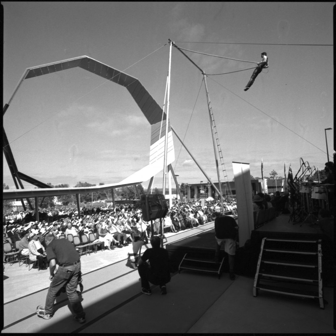 Black and white photograph shows a trapeze artist in midair in front of a seated audience