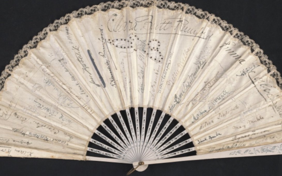 A handheld fan is spread open on a black backdrop; the fan has been signed by many different people