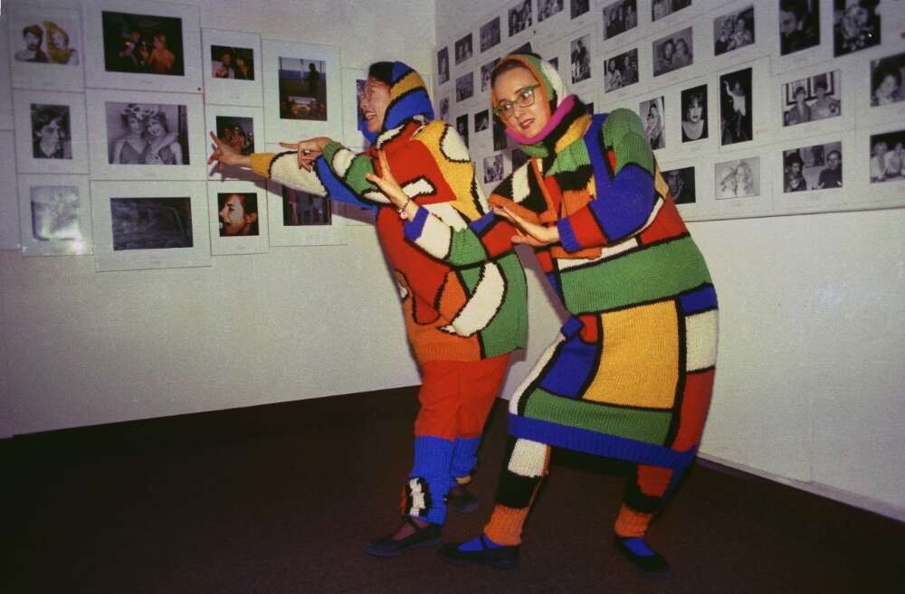 Two people dressed in bright coloured knitted garments pose in front of a wall of photos