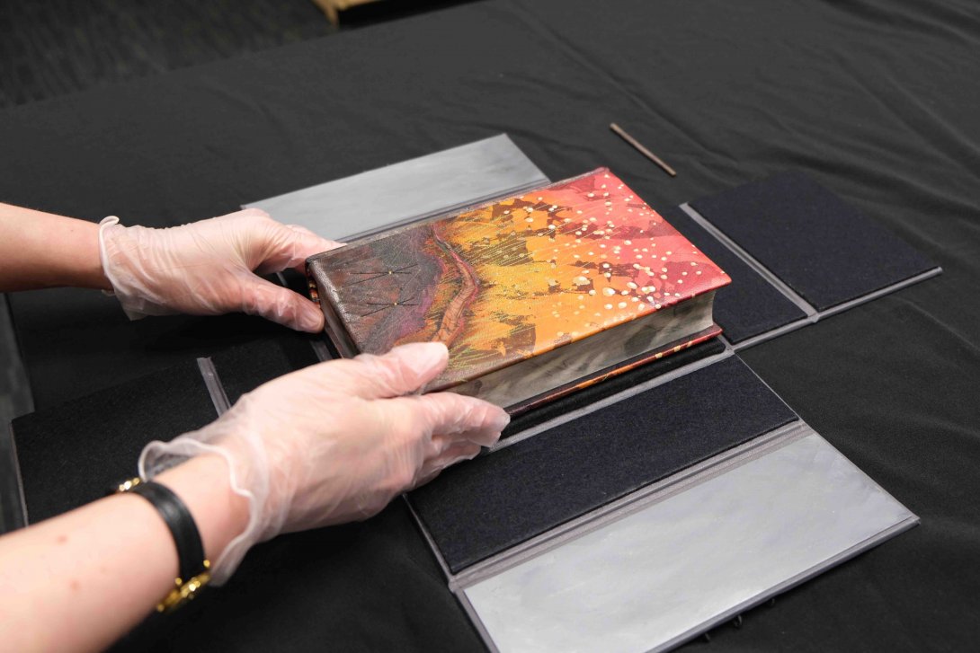 A painted hardback book is being placed into a black box by a pair of gloved hands
