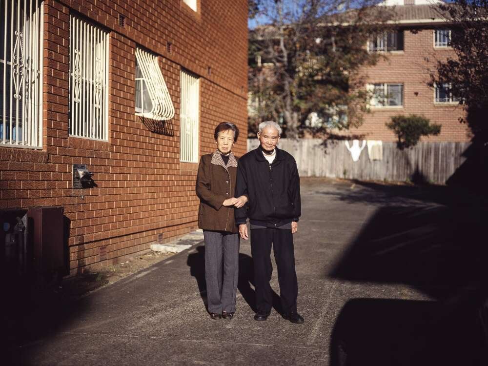 An elderly couple stand arm in arm outside a brick building