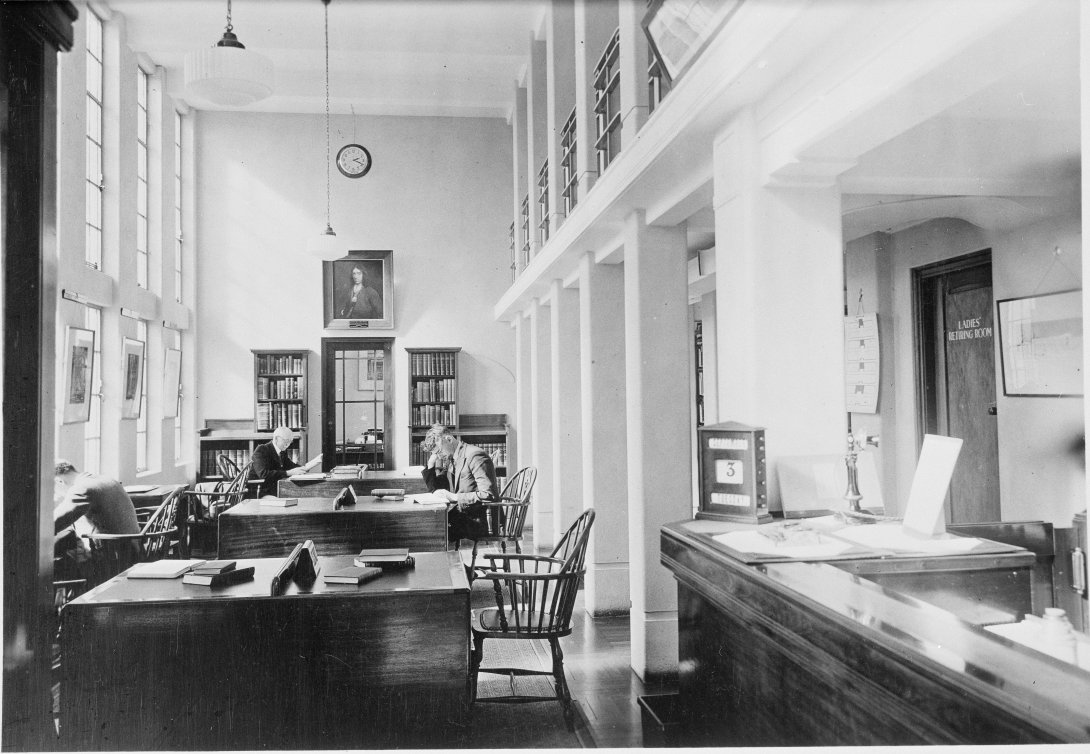 Black and white landscape of a small study area with desks and chairs through the centre, large windows to the right, a front desk with pillars behind it to the left 