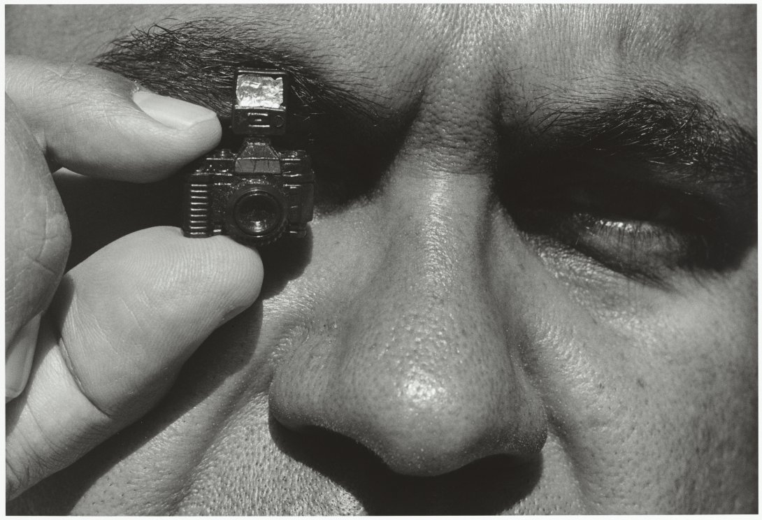A black and white close up of an Aboriginal man's face from above the brows to just below the nose. He holds a small camera over his left eye.