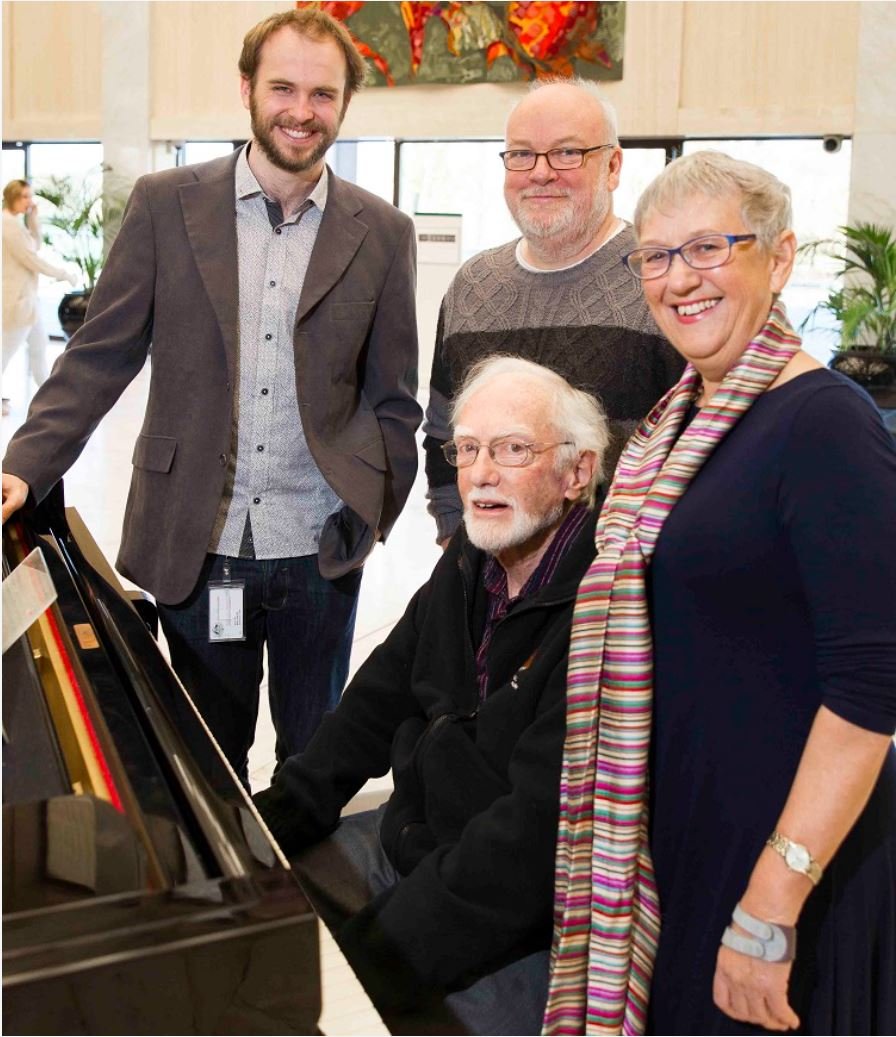 One person sitting at a grand piano, surrounding by 3 other people standing.
