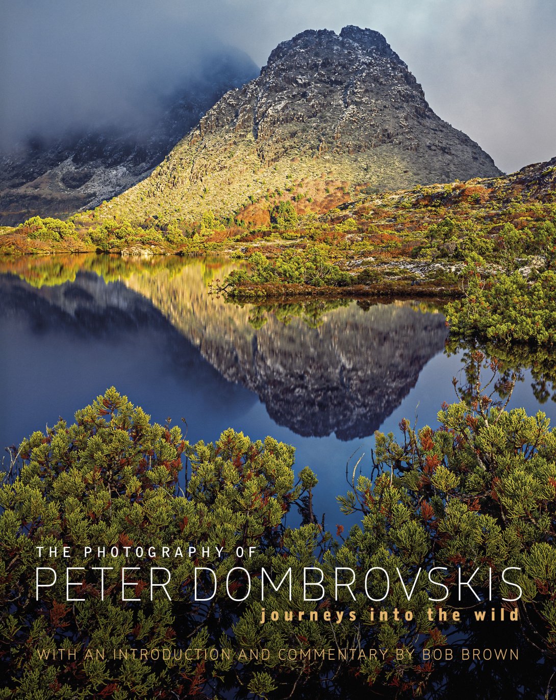 The Photography of Peter Dombrovskis, Journeys Into the Wild book cover. An image of a mountain reflected into still water with bushes at the base. 