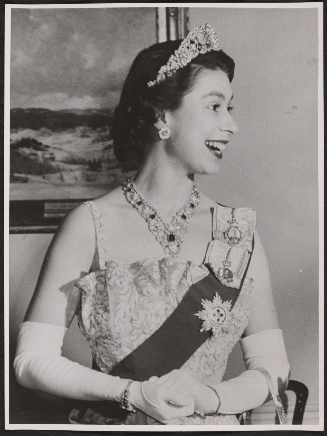 A black and white image of the Queen in 1954 dressed in full formal dinner attire, looking off to the side and smiling.