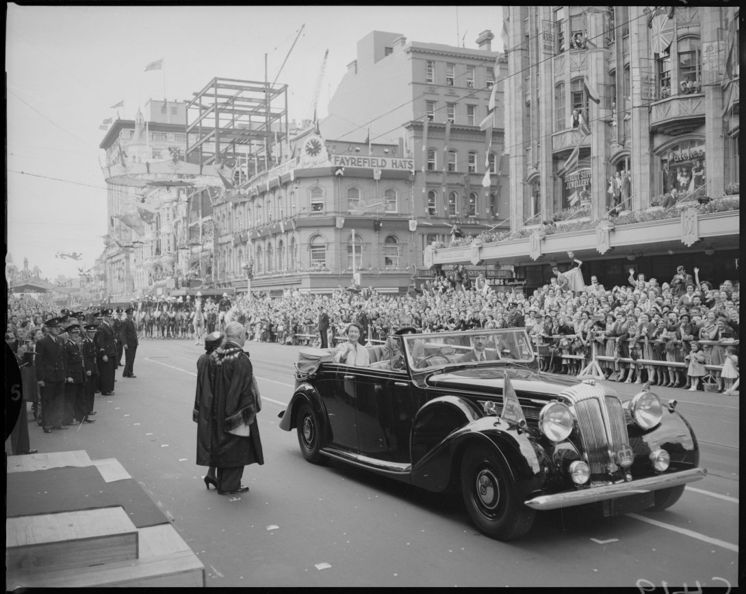 A black and white image of an open top car on the road. There are many people lining the road and tall buildings in the background behind the people.