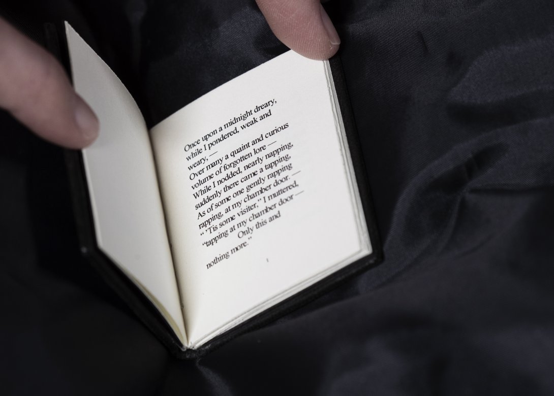 A miniature book being opened with fingertips with the start of 'The Raven' poem printed inside.