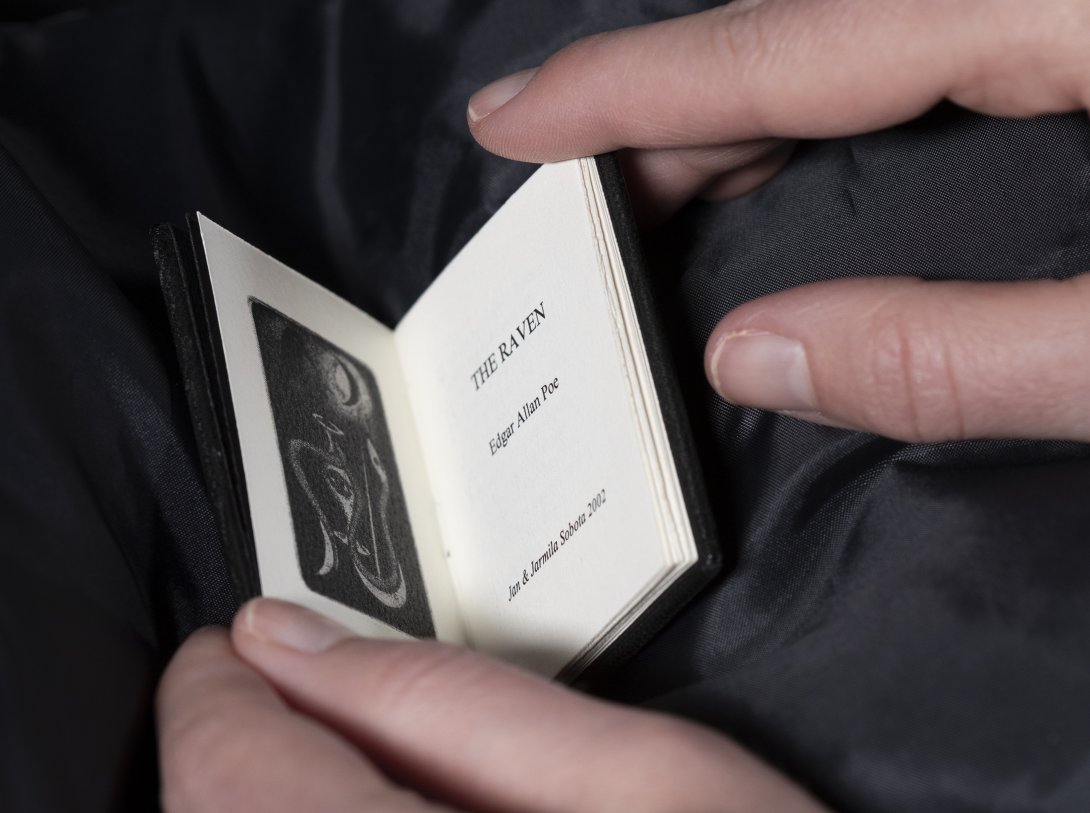 A miniature book being opened with fingertips. On the left is a sketched image of a snake looking p to the moon. On the right is the title page with text: 'The Raven, Edgar Allen Poe, Jan & Jarmila Sobota 2002'.