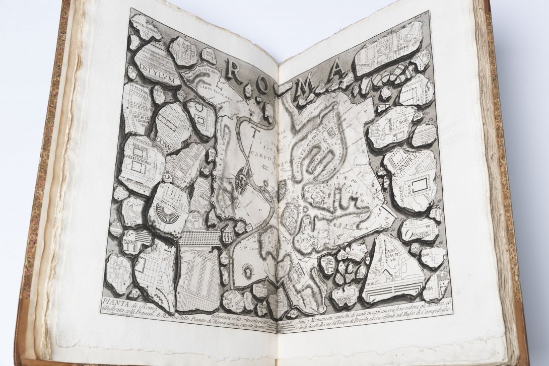 An old book open to show a two page spread of black and white engraved concrete with different illustrations and designs 'Roma' sits at the top