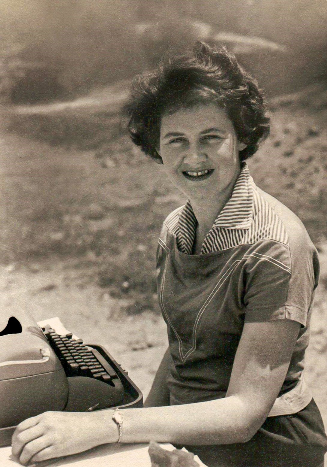 A sepia image of a woman looking at the camera. She is standing in front of a typewriter and is outside. 