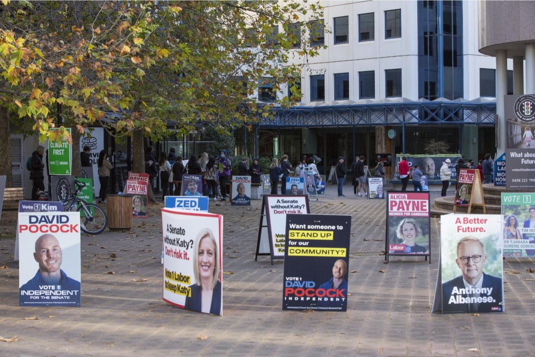 A paved square in front of a building with a tree to the left and people queueing out of the building. There are lots of political candidate posters and a-frames all through the square.