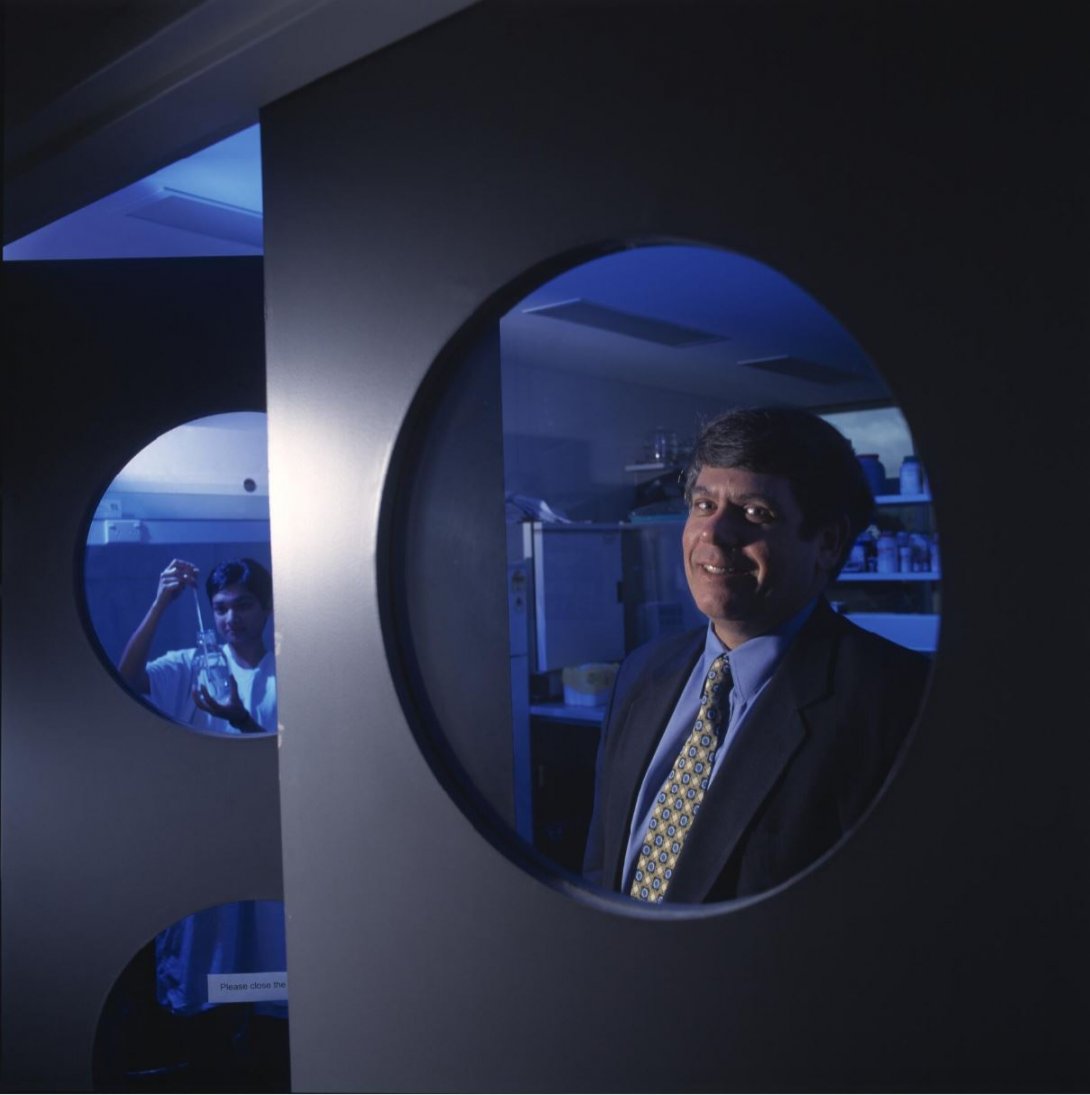 An image of a man standing behind a metal structure in a lab, with his upperbody appearing in a circular cut out in the structure. In the background is another person holding a long piece of lab equipment inside a jar. The whole lab is bather in a blue glow.