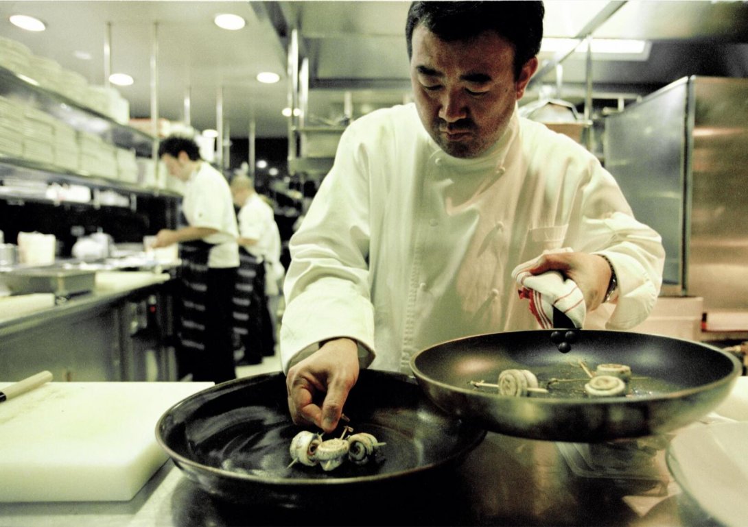A man in chef's whites in a commercial kitchen, plating up a dish. He is putting food from a frypan onto a plate. In the background there are 2 other chefs cooking food.