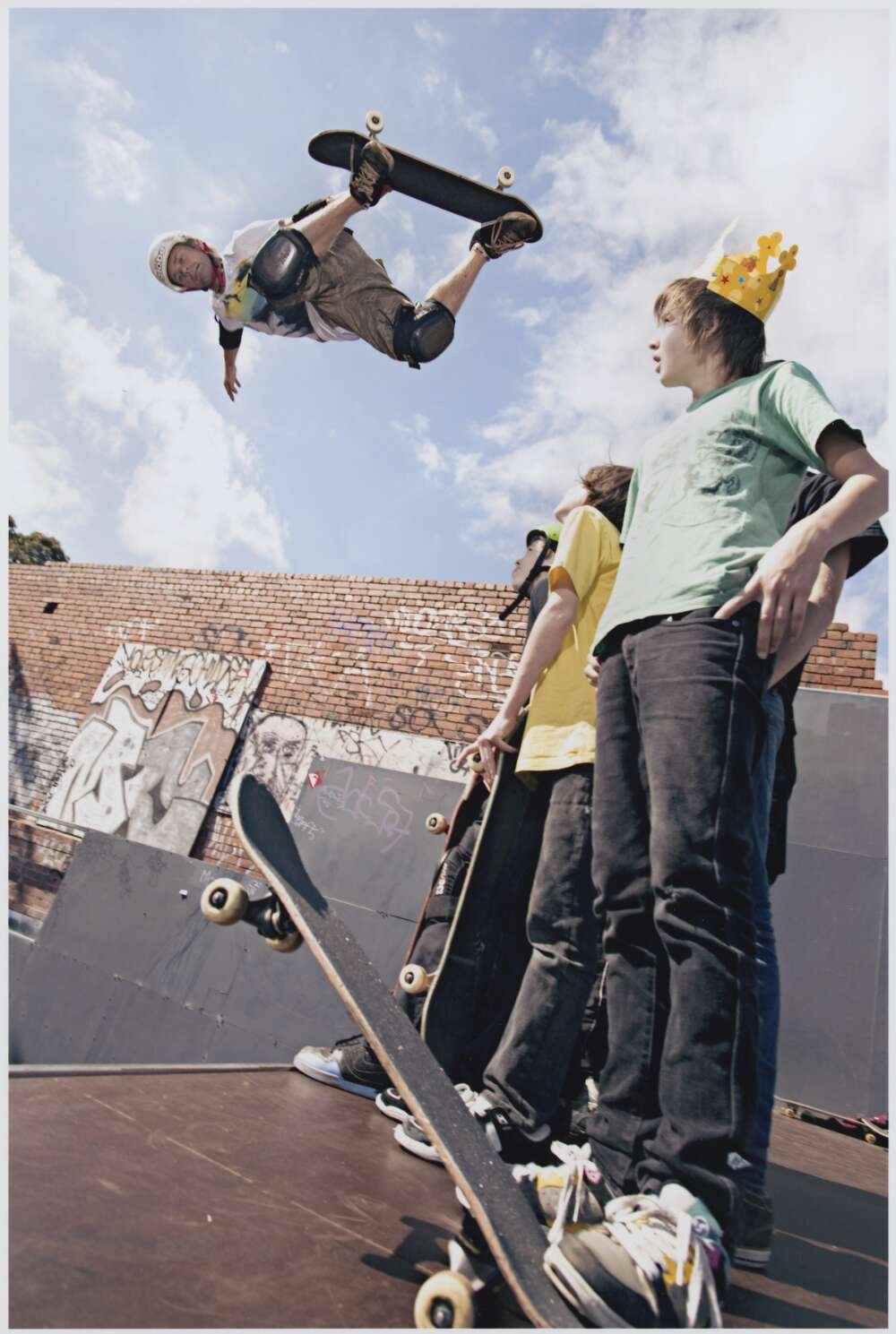 Several young skateboarders - one wearing a cardboard crown - look up at Renton Millar performing an aerial skateboard trick.