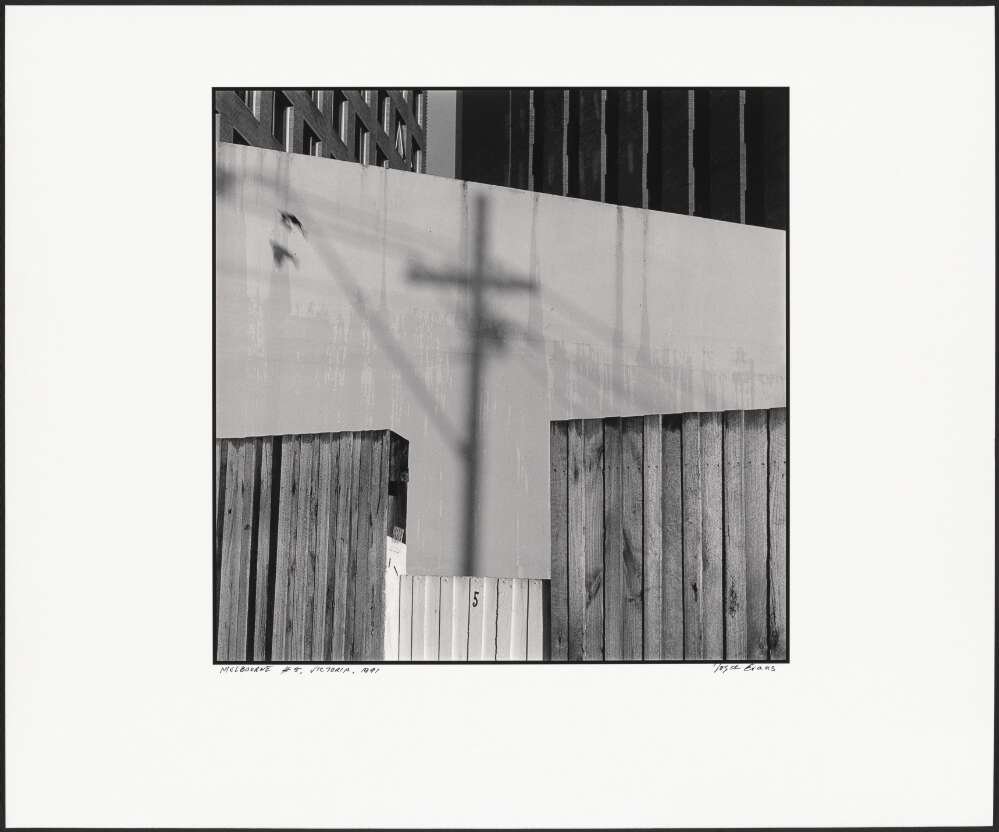 A fence with wooden planks at the bottom and some reflective metal on top. The metal reflects a powerline and an iron fence. Black and white.