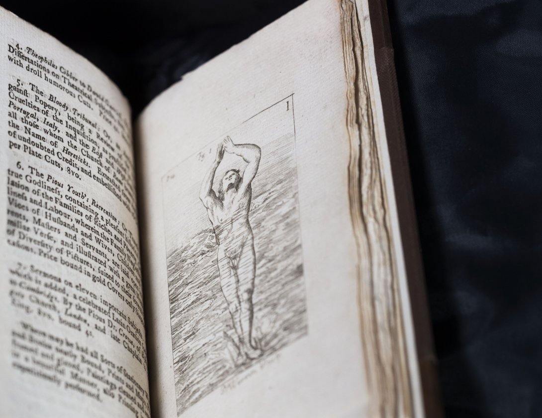 A rare book about swimming held open. Left side shows text too small to read, the right shows a black and white illustration of a man with his hands held together above his head, standing in waist-high water. 
