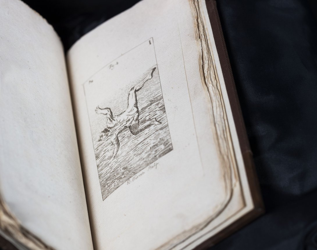 A rare book about swimming held open. Left side is a blank page while the right shows a black and white illustration of a man in water in a frog like pose. 