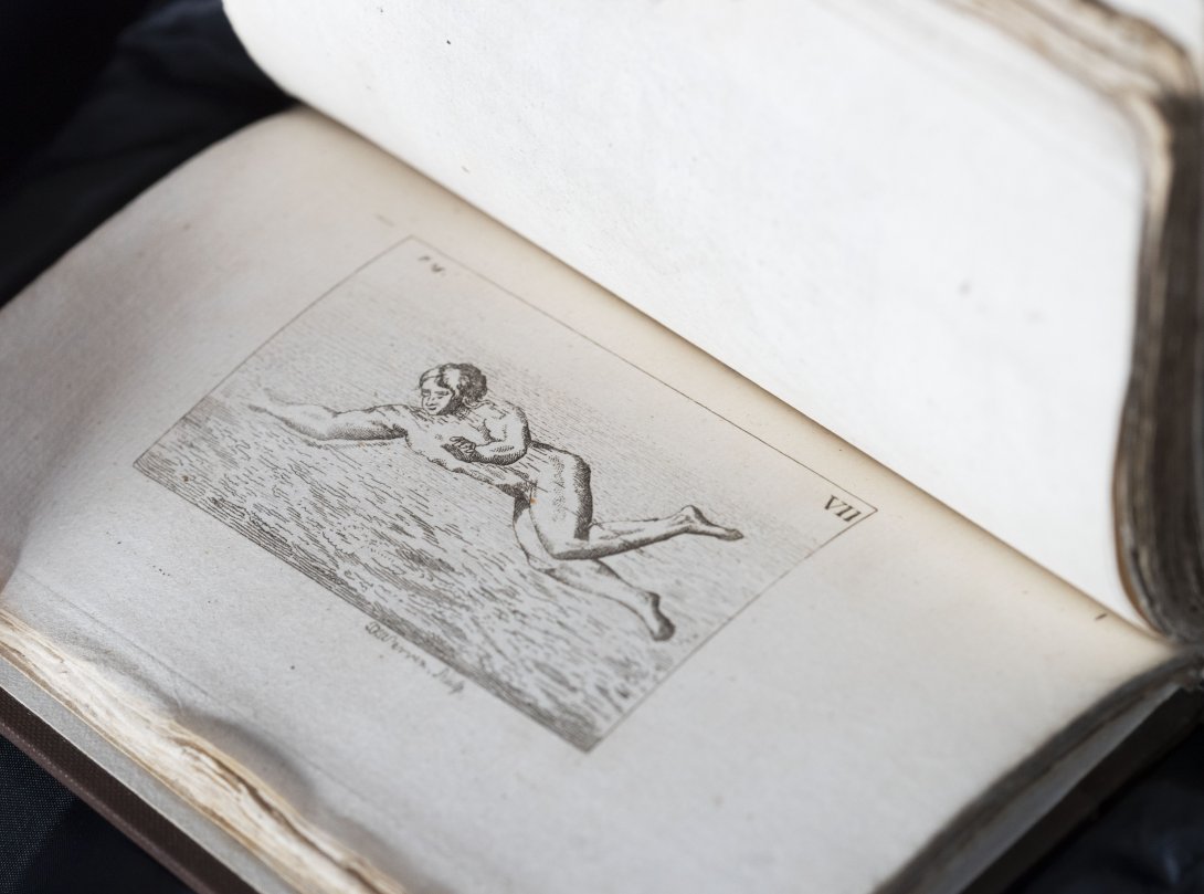 A rare book about swimming held open. Left side is an illustrated black and white person swimming on the surface of the water. 