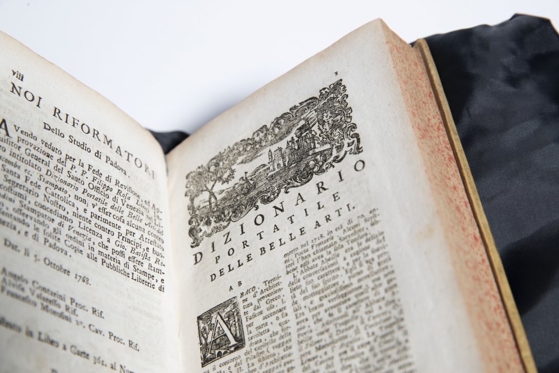 An old rare dictionary opened to a spread of pages with lots of text. On the right page, at the top, is an illustration of some buildings and nature framing it.
