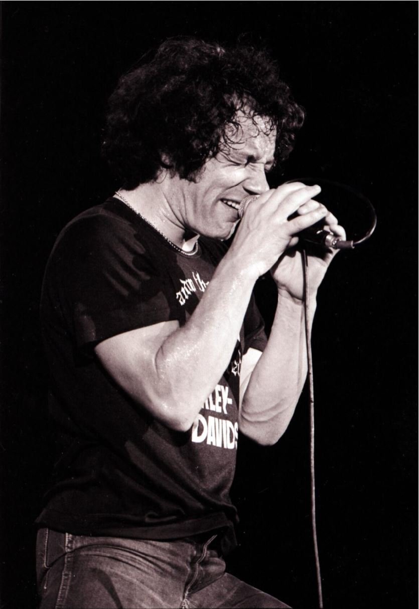 A black and white image of a man holding a microphone on a stand with 2 hands. He is holding the mic close to his mouth and has his eyes shut. He is wearing a black t-shirt and jeans.