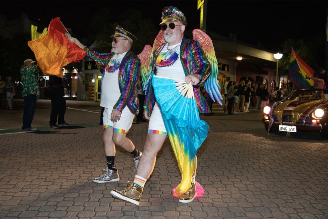 Two people marching in a parade at night time. They are dressed in colourful clothing and accessories.