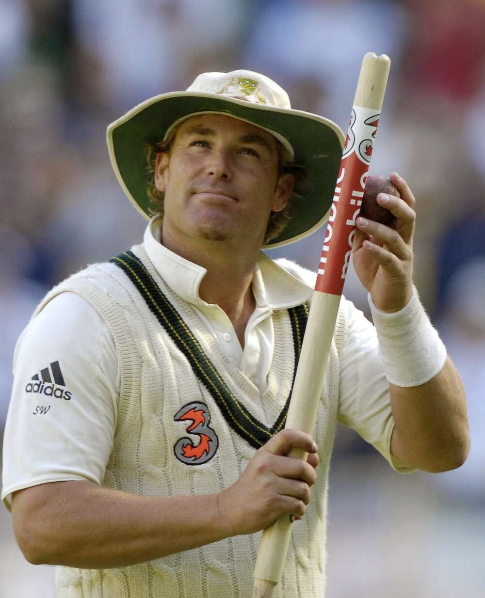 A photo of Shane Warne, dressed in test cricket whites and a hat, looking up at the crowd while holding a cricket ball against a stump he is holding in his other hand.