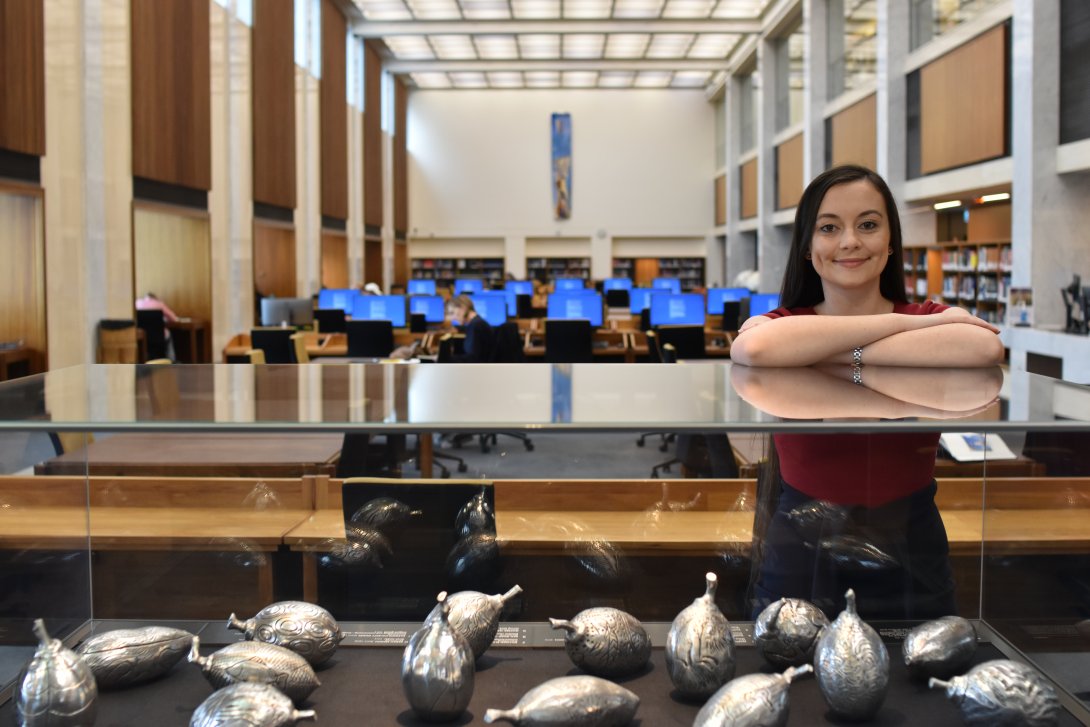 National Library of Australia 2022 scholarship recipient, Madeleine Pugin, stands smiling behind a glass cabinet containing Indigenous aluminium cast artworks by artists from Waringarri  Aboriginal Arts.