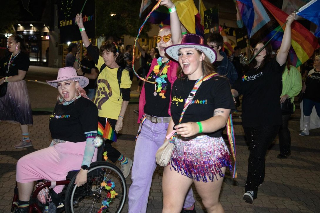 Several people marching in a parade at night time. They are wearing colourful clothes and accessories and some are waving rainbow and pride flags. One of the people is in a wheelchair.