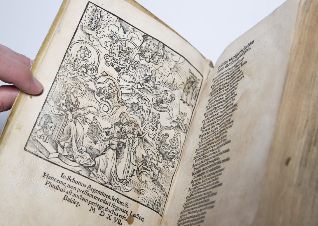 A rare book opened to show an detailed illustration  people, angels and ribbons to the left, a lift of words to the right. 
