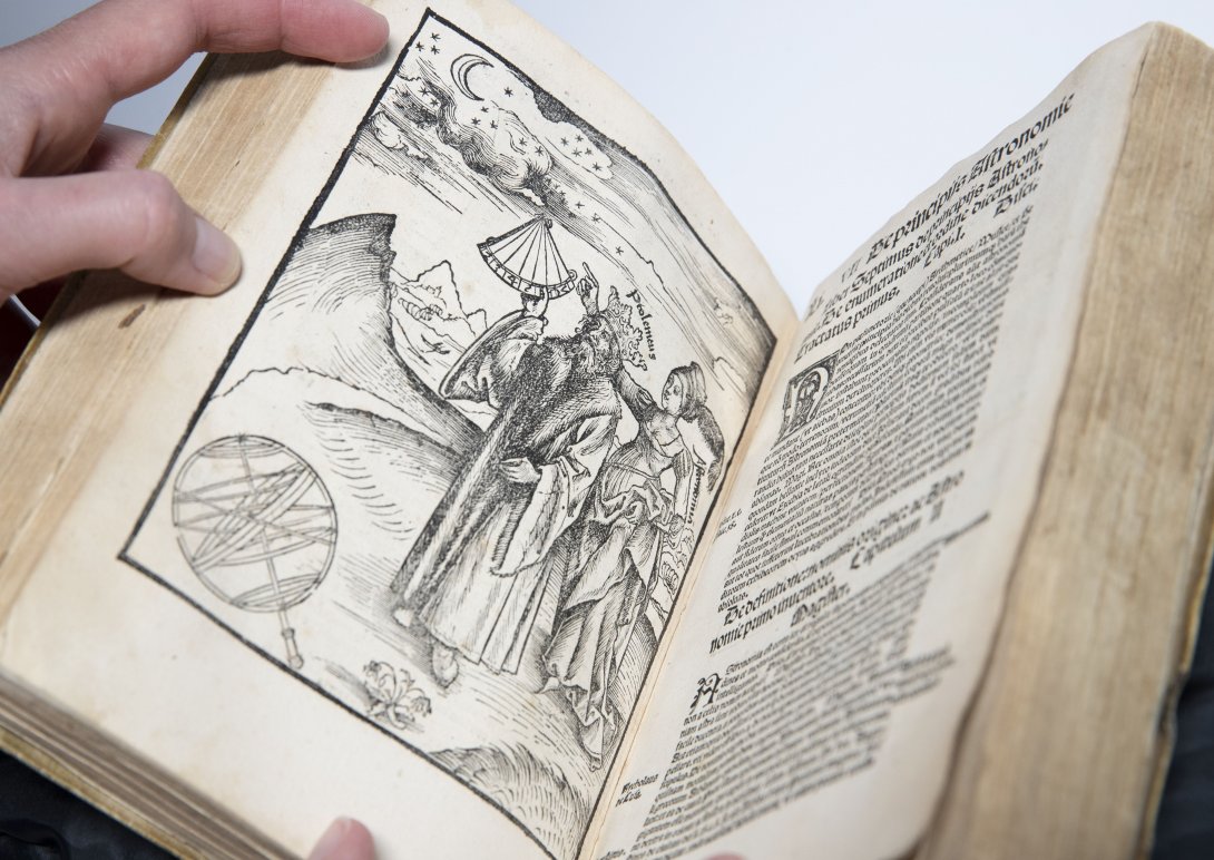 A rare book opened up to a detailed illustration of two people walking on mountainous fields on the left and a page of text on the left. 