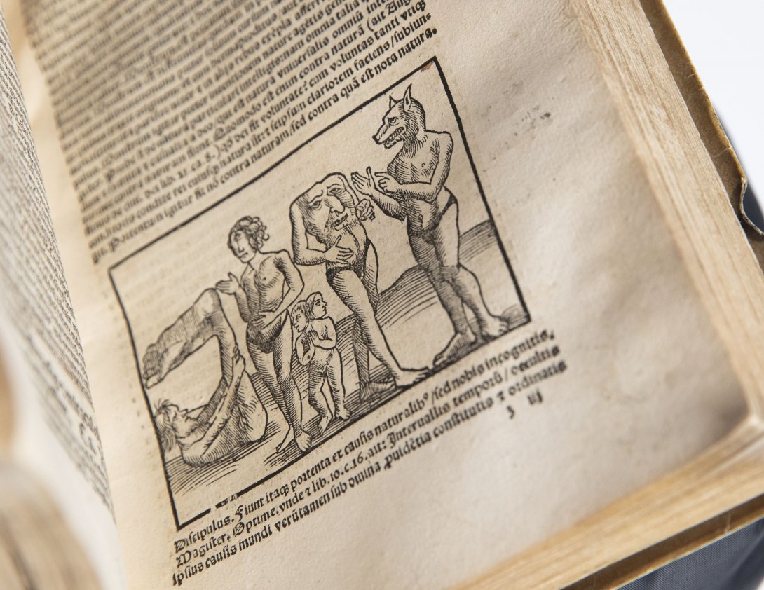 A rare book open to show an illustration of people with unusual features including one with a wolf head and another with a face on their chest. 