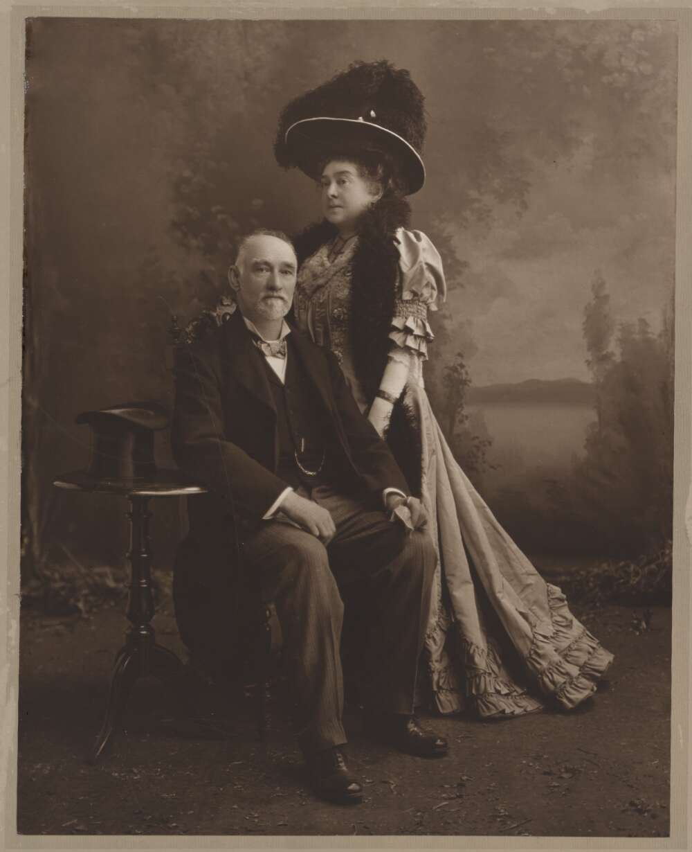 Woman in elaborate dress and large hat standing behind formally dressed man sitting on a chair