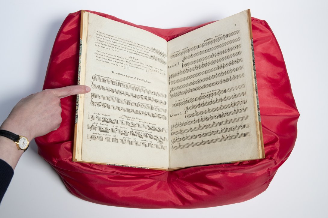 An open music textbook sitting on a red cushion with a woman's right hand holding the page open to the left.