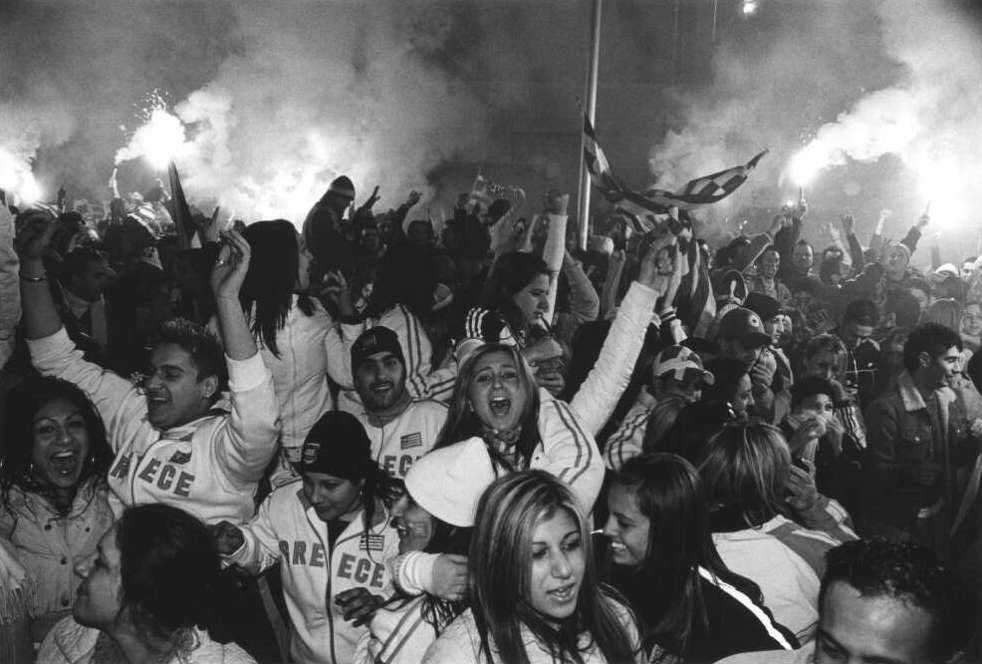 A large crowd celebrate at night time. Smoke fills the air as flares are lit and a Greek flag is raised. Many of the supporters are wearing the Greece's soccer team merchandise