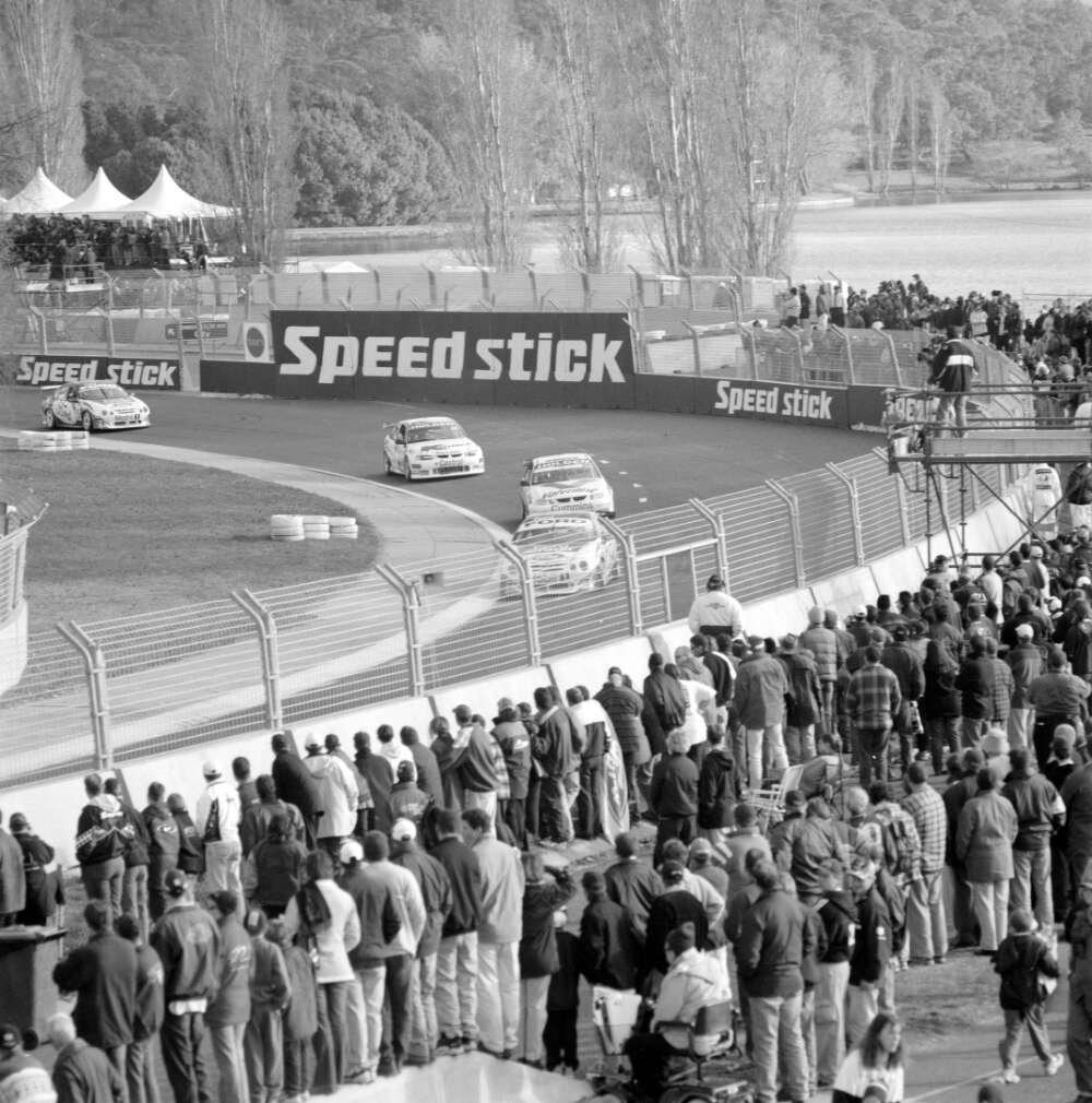 A crowd of onlookers watch from behind a fence as four race cars speed past. Lake Burley Griffin can be viewed in the background.