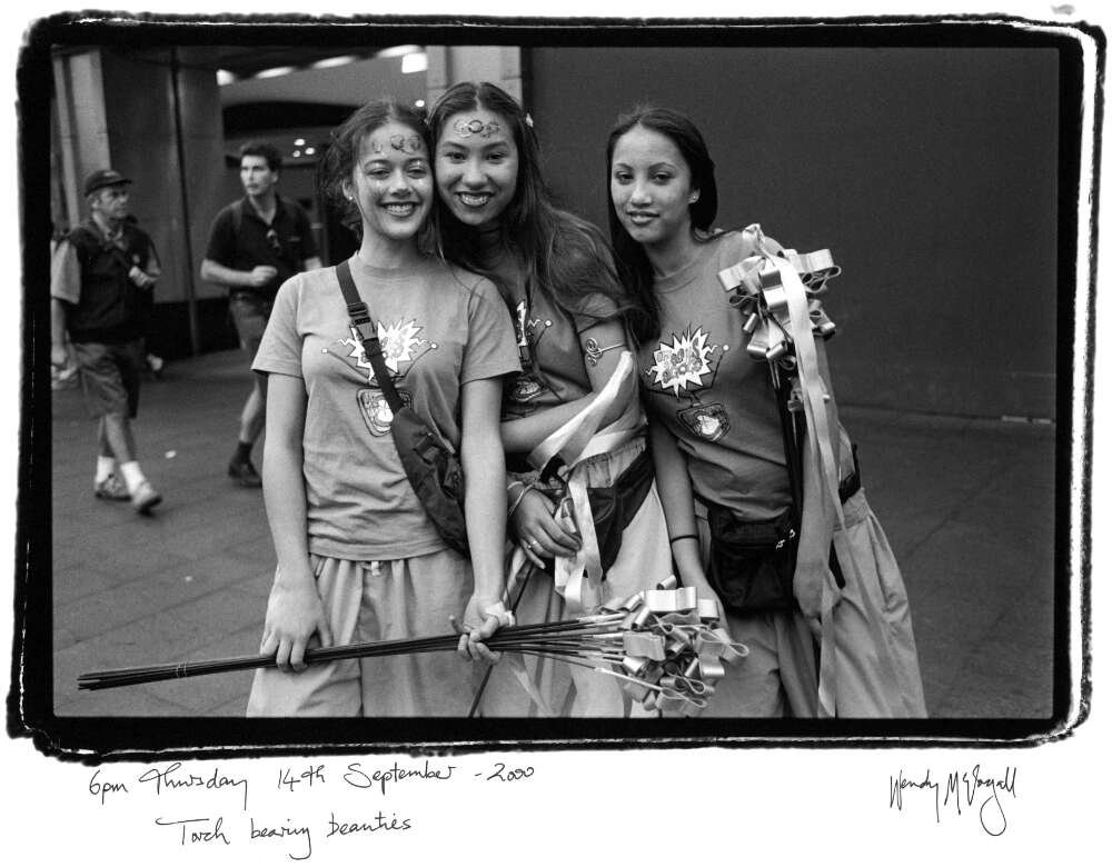 Three young women stand posed for the camera, smiling. Two of the women have olympic rings painted on their foreheads, and all three are wearing matching t-shirts. They are carrying ribbon batons for sale.
